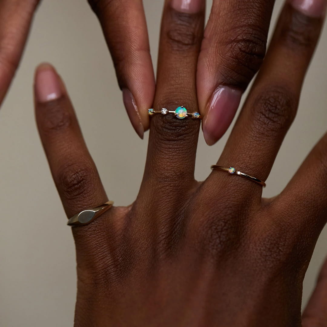 Model's hand wearing stacked rings, including an opal solitaire and a thin gold band, neutral background