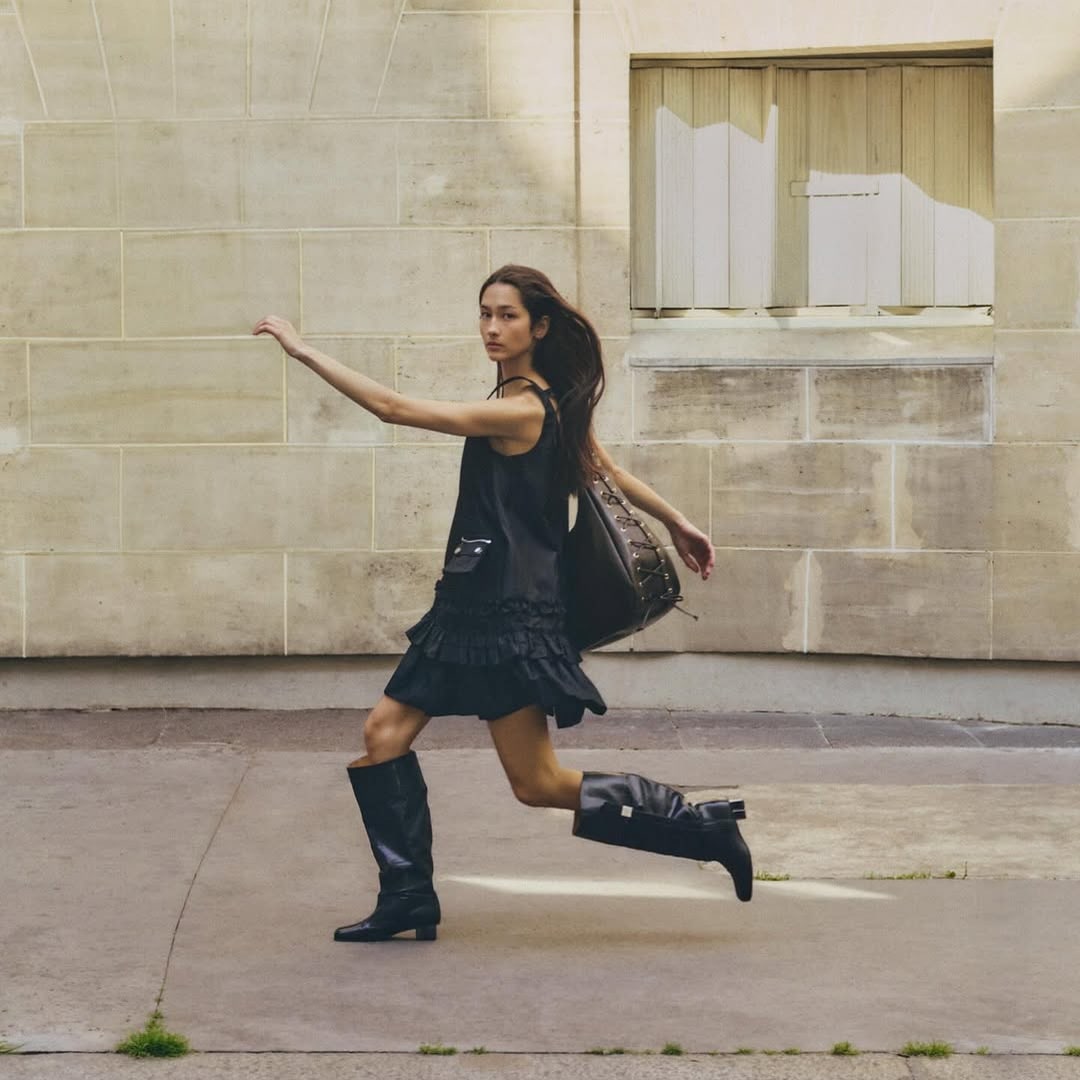 Woman in black dress and boots walking outdoors with a large bag.