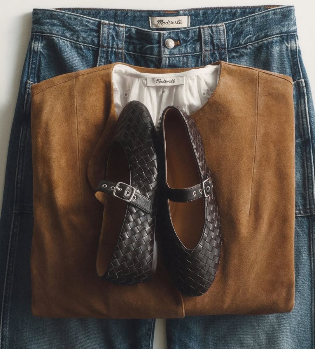 Black woven Mary Jane shoes placed on a brown suede jacket and blue jeans in a flat lay.